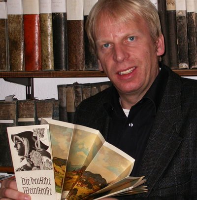 Photographie von Christof Krierger vor einem Bücherregla, einen auseinandergefalteten Flyer zur Deutschen Weinstraße in der Hand.