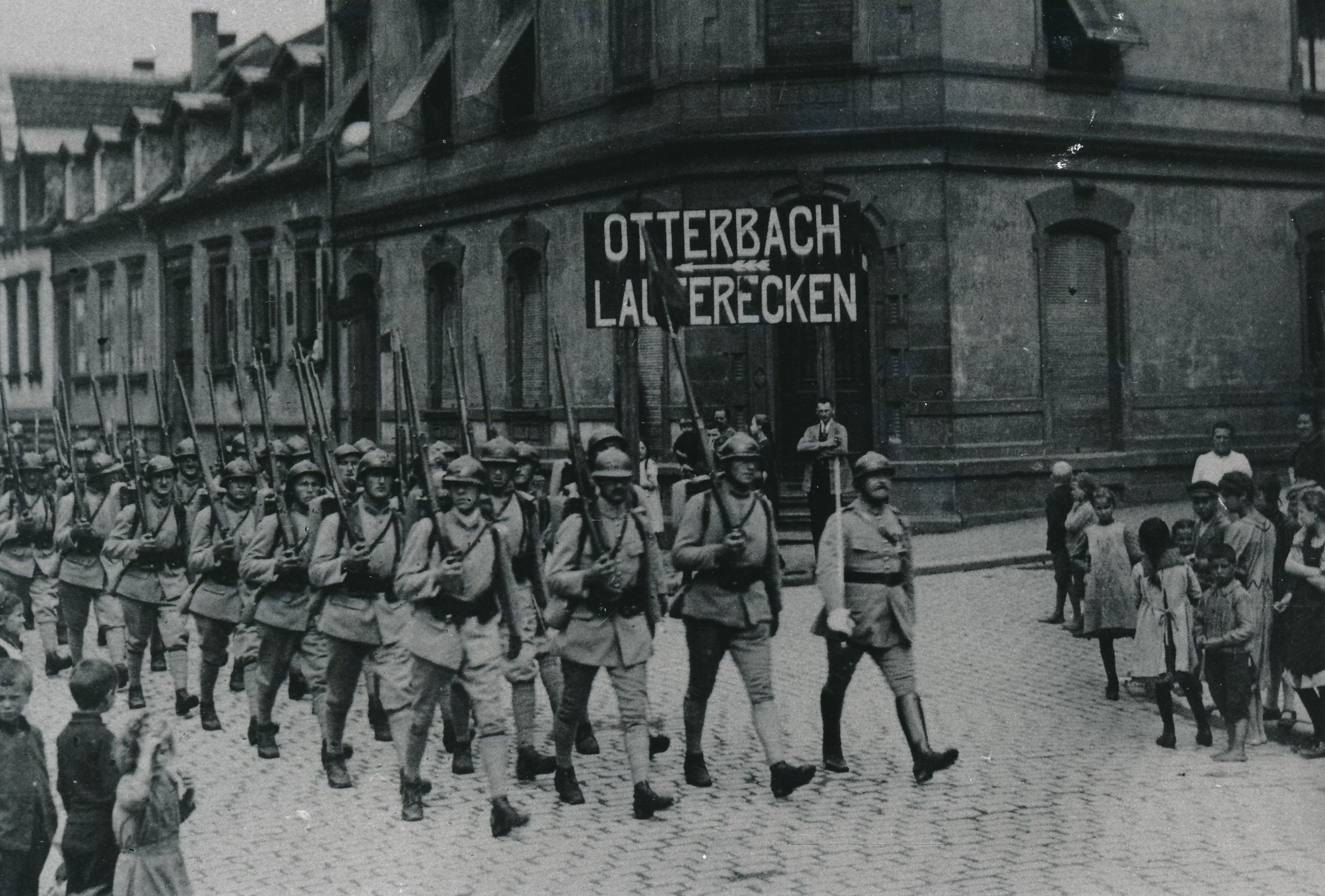 Eine Kolonne französischer Soldaten, jeweils 4 pro Reihe, marschiert über eine Straße, beobachtet von Kindern. Zwei der Soldaten in der ersten Reihe halten ein Schild auf dem Otterbach steht.