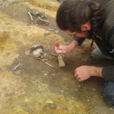 Ein Archäologie kniet auf dem Boden und legt mit einem Pinsel ein Skelett frei.