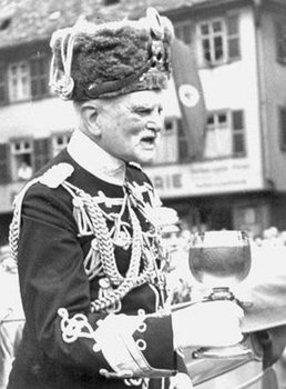 Bild des alten August von Mackensen in Uniform mit zahlreichen Orden, ein übergroßes Glas in der Hand haltend.