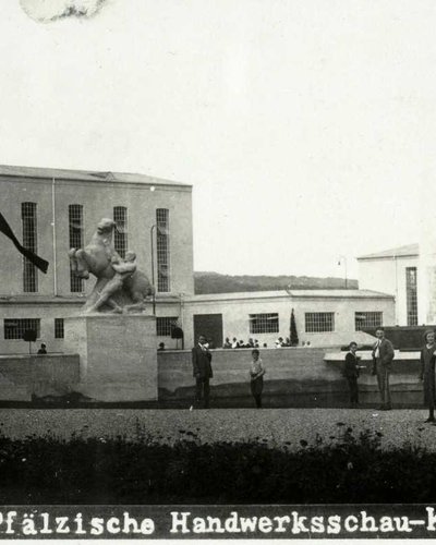 SW-Foto ›Pfälzische Handwerksschau Kaiserslautern 1925‹. Hintergrund: moderne weiße Gebäude im Bauhaus-Stil. Mitte: ein Wasserbassin, gerahmt von zwei weißen Rossbändiger-Figuren. Um das Bassin: viele Besucher.