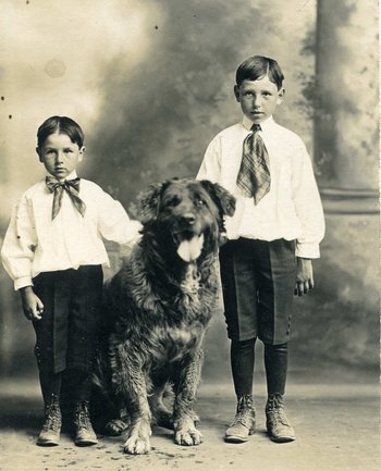 Auf der schwarzweißen Fotografie stehen Jungen links und rechts neben einem zotteligen Hund. Beide Jungen (verm. 5–8 Jahre) tragen ein weißes Hemd, eine Schleife um den Hals, knielange, dunkle Hosen, darunter lange Strümpfe und zeitgenössiche Lederschuhe.