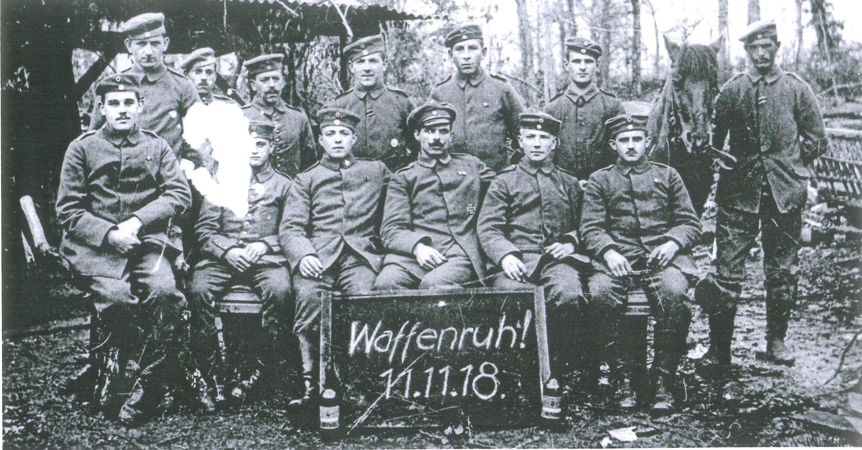 Soldaten in zwei Reihen aufgereiht für ein Foto. Die erste Reihe sitzt, die hintere steht. Einer der hinteren Soldaten hält ein Pferd am Zügel.  Vor der vorderen Reihe steht eine Tafel worauf mit Kreide geschrieben steht, Waffenruh!, 11.11.18.