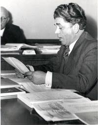 Photographie von Willy Feller an einem Tisch sitzend eine Text vorlesend, vor ihm zahlreiche Texte und aufgeschlagene Bücher. Im Hintergrund an einem anderen Tisch ein Mann in Anwaltsrobe.