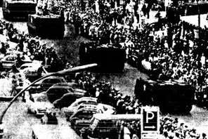 Photographie von Panzer die über eine Straße fahren, umringt von einer Menschenmenge. Hinter den Menschen auf der linken Seite des Bildes parken zahlreiche Autos.