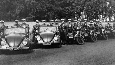 Schwarzt-weiß Photographie zahlreicher Polizisten aufgereiht in zwei VW Käfern und mehreren Motorrädern mit Beiwagen.