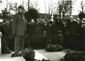 Photographie von August Beckers Grab, daneben steht ein Mann in Uniform und mit Pickelhaube, hinter ihm eine Versammlung von Trauergästen.